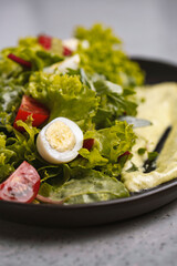Fresh green salad with quail eggs, cherry tomatoes and guacamole in black plate on grey background