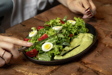 Person eating Fresh green salad with quail eggs, cherry tomatoes and guacamole in black plate on grey background