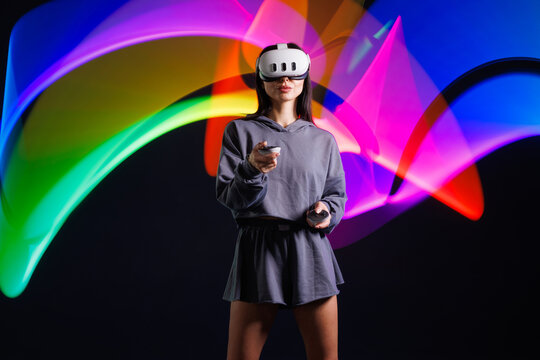 Young woman in VR headset playing virtual reality game with motion controllers, neon light trails in futuristic immersive environment.