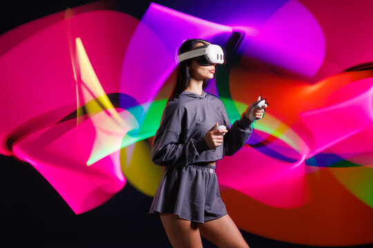 Young woman in VR headset playing virtual reality game with motion controllers, neon light trails in futuristic immersive environment.
