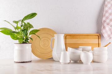 Beautiful kitchen background with a set of white ceramic utensils, wooden cutting boards, green houseplant in a stylish pot. Front view.