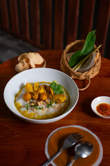 Bubur ayam, a traditional Indonesian food. Rice porridge with sliced chicken, rolled omelette, peanut, sambal (chili sauce), and kerupuk (crackers), poured with curry sauce. Served on wooden table