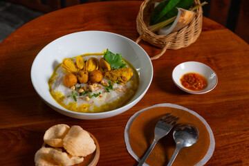 Bubur ayam, a traditional Indonesian food. Rice porridge with sliced chicken, rolled omelette, peanut, sambal (chili sauce), and kerupuk (crackers), poured with curry sauce. Served on wooden table