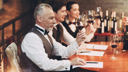 Three sommeliers assess taste and color qualities of wine when examining glass in restaurant.