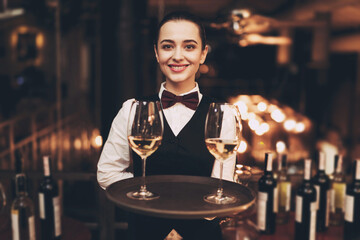 Joyful waitress holds tray with glasses of white wine, standing near bar counter.