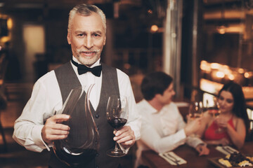 Sommelier is holding glass of wine and decanter, standing near table of couple who are on date.
