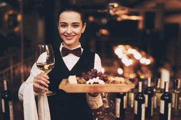 Joyful elegant waitress holding glass of white wine, plate of cheese and fruit, standing near bar.