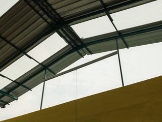 Industrial metal roof with transparent polycarbonate panels and a yellow wall, showing an open ventilation section. Structural steel framework visible against a cloudy sky.