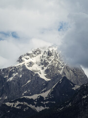 Obraz premium A majestic snow-covered mountain peak partially hidden by thick clouds in the Friuli Venezia Giulia region, Italy. The rugged rock formations contrast with the soft, billowing clouds.