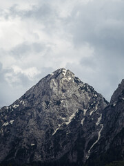 A sharp, rocky mountain peak with patches of snow, set against a dramatic cloudy sky in the Friuli Venezia Giulia region, Italy. The rugged terrain highlights the raw beauty of the Alps.
