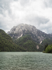 A tranquil view of Lago del Predil in Italy, surrounded by lush green mountains and towering rocky peaks. The calm turquoise waters reflect the dramatic cloudy sky above.
