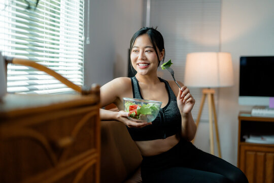 Fit young Asian woman in athletic wear savoring fresh vegetable salad, resting on living room sofa post-workout, radiating wellness and vitality