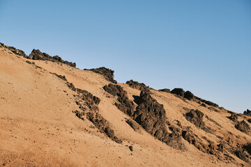 Dramatic landscape photo of a sandy hillside with dark volcanic rocks under a clear blue sky. ...