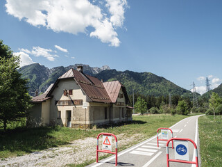 An abandoned railway station along the Alpe Adria cycling route near Tarvisio, Italy, with a mountain backdrop, red barriers, and power lines under a bright blue sky.