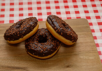 Three chocolate glazed donuts topped with sprinkles are arranged neatly on a wooden board. The background features a classic red and white checkered tablecloth