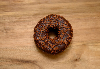 A freshly baked chocolate donut adorned with crunchy chocolate sprinkles rests on a wooden surface, showcasing its glossy glaze and inviting appearance