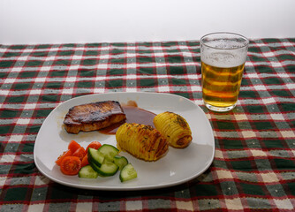 A delicious plate with grilled pork chop, potato, and fresh cucumber and tomato slices sits on a checkered tablecloth next to a cold drink, ready to be enjoyed