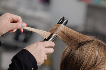 Professional hairstylist curling blonde hair with flat iron in beauty salon, close-up of styling process and haircare techniques
