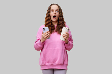 A young woman with wavy hair expresses surprise while holding a smartphone in one hand and a coffee...