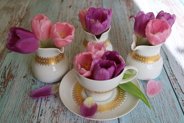 On the wooden table, there is a cup filled with purple tulip flowers and three milk jugs with...