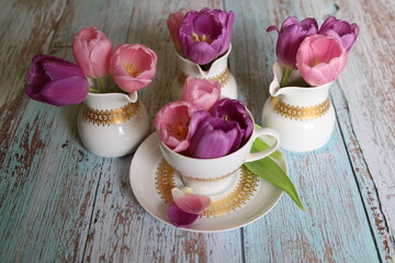 On the wooden table, there is a cup filled with purple tulip flowers and three milk jugs with tulips.  Easter Table Decoration. Springtime decor. 