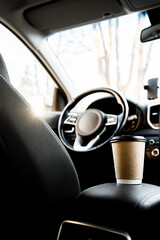 A recyclable coffee cup and lid are on the armrest next to the driver in the car interior, view...