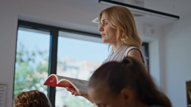 Woman teacher dictating lesson to pupils walking with book at classroom closeup.