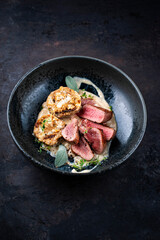 Roasted fillet of saddle of venison with bread dumplings, mushrooms and bean sticks served with lingonberry sauce as close-up on a design plate with a rustic background
