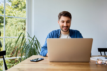 Happy young man working on laptop at home sitting at table