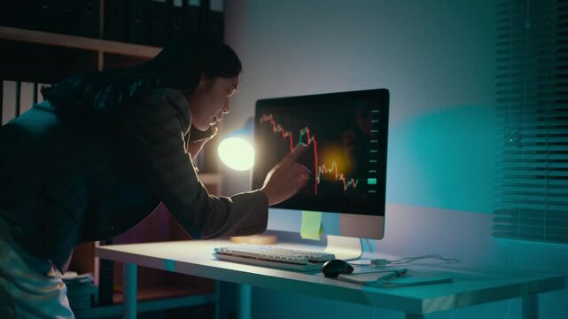 Professional female analyzing downward financial graph on computer screen, working late with desk lamp illuminating dimly lit office environment