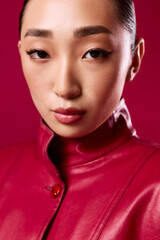 Stylish asian woman in red leather jacket posing with hands on hips against red background, looking to the side