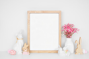 Modern Easter home decor shelf scene. Mock up portrait wood frame with Easter bunnies, eggs and pink flowers on a white shelf. Copy space.