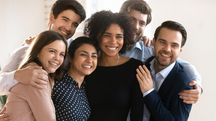 Six multiracial young and friendly teammates hugging standing together indoor, smile, looking away, exuding friendship, amity, racial equality, celebrate joint professional success. Teamwork, synergy
