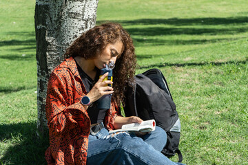 Young student drinking water and reading a book under a tree in a park