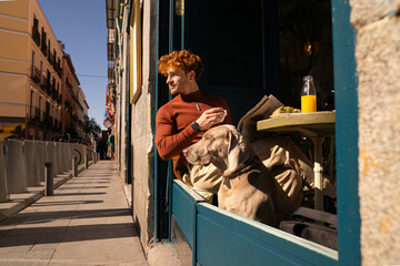 Sunny scene of Young ginger haired boy and his dog on the terrace of a cafe having a healthy breakfast
