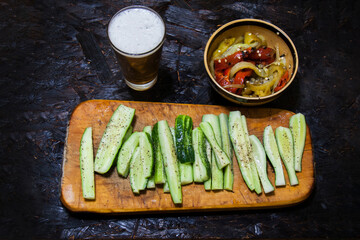 The photo shows a vegetarian snack with beer: sliced cucumbers, baked peppers and beer. Sliced cucumbers on a wooden board, baked peppers in a bowl and beer in a glass.