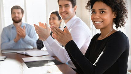 Obraz premium Multinational millennial audience participate in seminar or corporate training seated at conference table, applauding, clap hands, express appreciation to coach for presentation. Recognition
