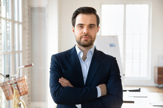 Head shot portrait confident businessman dressed in elegant formal suit posing with arms crossed in modern conference room, ready for negotiation, feels self-assured, satisfied with project completion