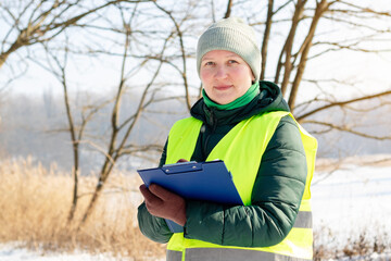 Female ecologist inspects trees in winter. Middle aged female volunteer with a tablet checks trees...