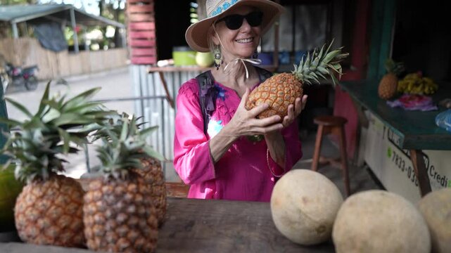 A happy woman smells a pineapple while exploring a local market in Moyogalpa on Ometepe Island, Nicaragua. Surrounding her are fresh fruits and vibrant scenery.