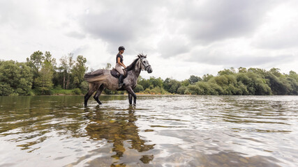 Gray horse with a female rider in the saddle walking through river water. Ecotourism and equestrian...