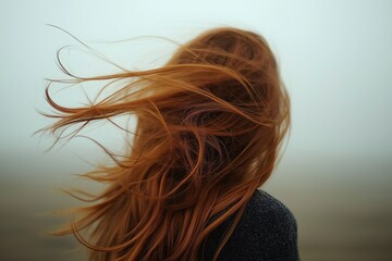 Back view of ginger woman with long, flowing red hair being blown by strong wind on foggy background.