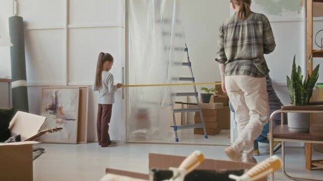 Adorable little girl in pants and pullover helping her father measure width of doorway of living room with tape in new apartment while young woman walking and taking notes in notepad
