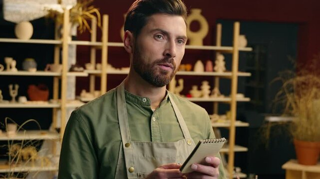 Happy cheerful Caucasian man male small business owner potter entrepreneur businessman writing notes notebook write thinking having idea eureka insight inspiration creativity indoors pottery workshop
