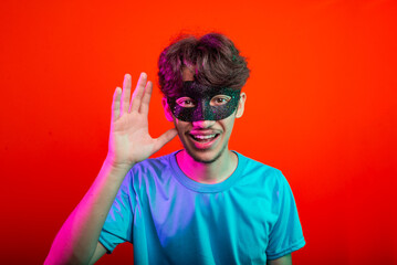 Happy funny handsome young man wearing carnival mask looking and making hand gestures to the camera. Isolated on neon orange background.