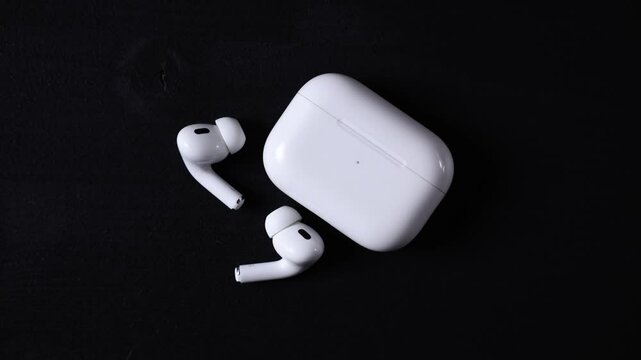 Pair of white wireless earbuds with matching charging case on a dark black surface, featuring compact design and LED indicator on the case.