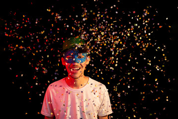 Fototapeta premium Young man in white shirt, wearing carnival mask and green hat having fun with paper confetti. Isolated on black background.