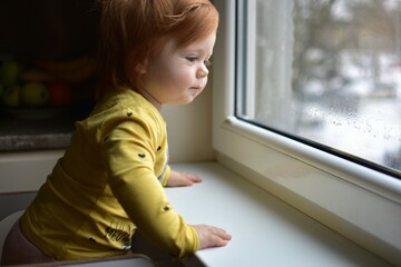Red messy hair toddler girl curiously looking through the window. Daughter impatiently waiting...