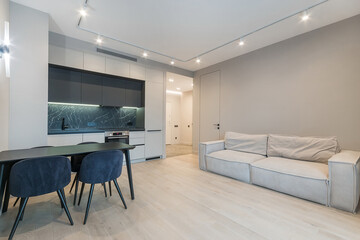 The bright decor of the studio apartment with beige laminate. A white kitchen set with black trim, a dining table and a light sofa.