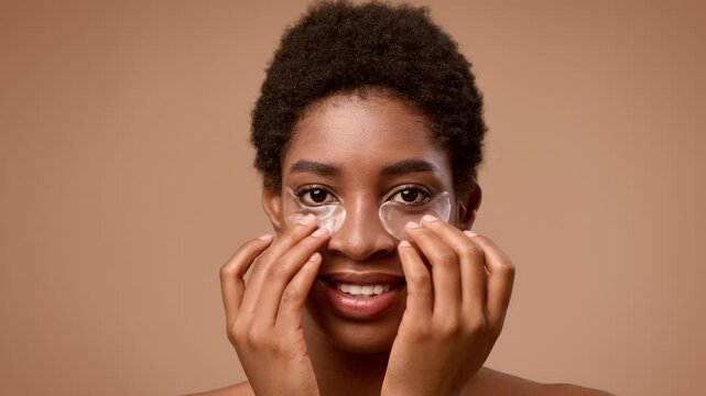A young, short-haired African American woman smiles while applying gel eye pads. She is half-dressed against a neutral background, showcasing her skincare self-care routine.